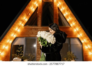 Woman holding a bouquet of white roses in front of a cozy wooden house illuminated with string lights at night - Powered by Shutterstock - Get 15% off with code: PIKWIZARD15
