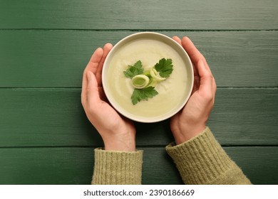 Woman holding bowl of delicious leek soup at green wooden table, top view