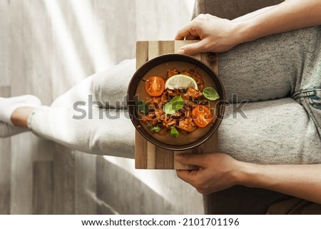 Woman is holding a bowl of thick tasty lentil soup with fresh vegetables on his knees.