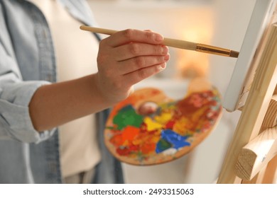 Woman holding brush and palette painting on easel with canvas indoors, closeup - Powered by Shutterstock