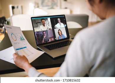 Woman Holding A Business Report Working At Home Having A Video Conference With Colleagues. Over The Shoulder View Of A Businesswoman Having Online Business Meeting From Home.