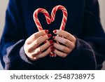 Woman holding Christmas candy canes, close up. Heart shape made of two candy canes. Charity concept, female holds two candies forming heart at chest level. Sweet caramel for children. Cozy sweater.