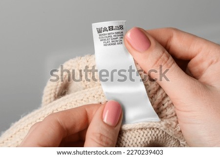Woman holding clothing label on beige garment against light grey background, closeup