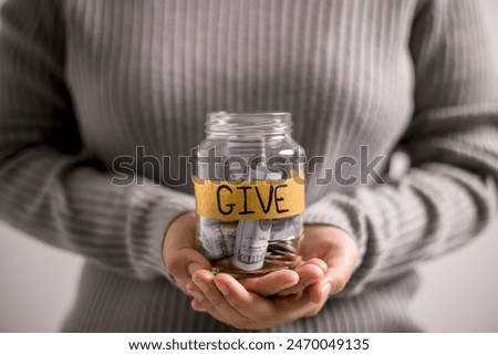 Woman holding coins and cash in jar with words Give or Donate elegantly lettered on its front, embodying spirit of giving. words, lettering, donate, jar, money, giving, woman, give, coin.