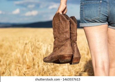 Woman Holding Cowboy Boots In Field. Cowgirl Walking In Countryside. Worn Leather Riding Boot