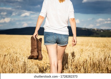 Woman Holding Cowboy Boots In Field At Summer. Cowgirl Walking In Countryside. Worn Leather Riding Boot