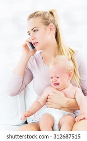 Woman Holding Crying Baby Talking On Mobile Phone
