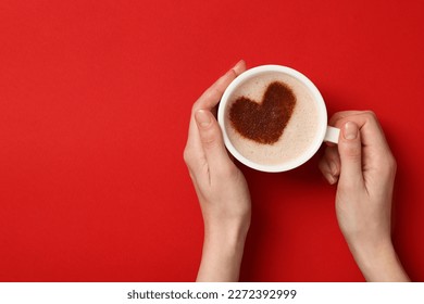 Woman holding cup of aromatic coffee with heart shaped decoration on red background, top view and space for text