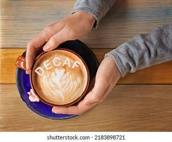 Woman holding cup of aromatic decaf coffee with foam at wooden table, top view