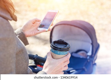 Woman Holding Cup Of Coffee To Go And Using Smartphone During Walk With Baby Carriage. Modern Young Business Woman Combining Work And Child Care