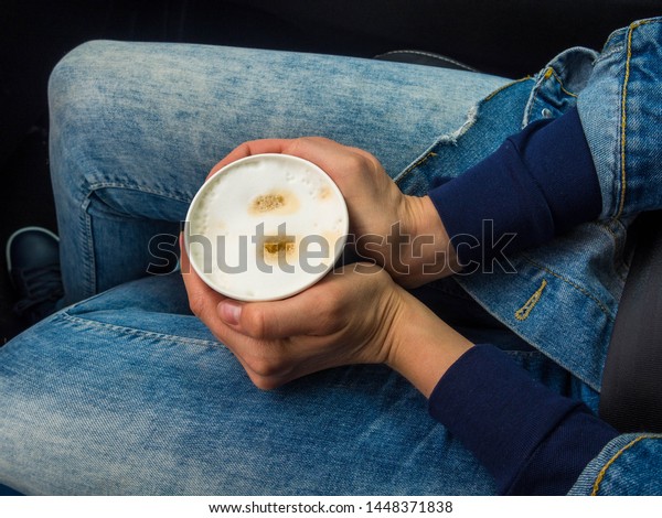 Woman is holding cup of coffee inside of car.\
Travel lifestyle. Legs in\
jeans