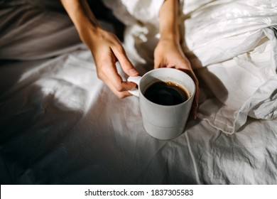 Woman Holding A Cup Of Coffee, Sitting On Bed In The Morning Light.