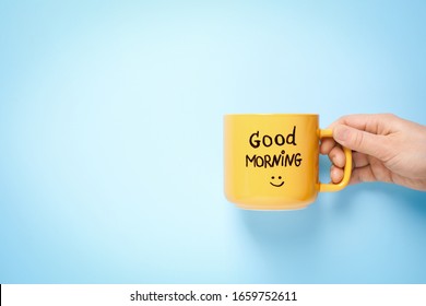 Woman holding cup of coffee with words GOOD MORNING on light blue background, closeup. Space for text
