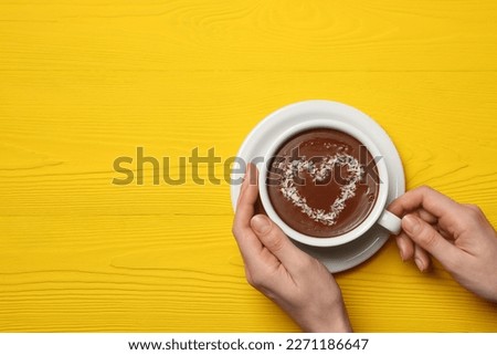 Woman holding cup of hot chocolate with heart shaped decoration at yellow wooden table, top view. Space for text
