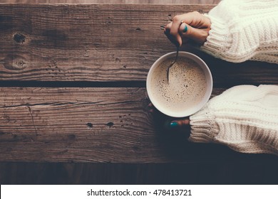 Woman holding cup of hot coffee on rustic wooden table, closeup photo of hands in warm sweater with mug, winter morning concept, top view