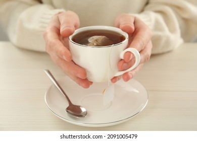 Woman holding cup with tea bag and hot water at white wooden table, closeup