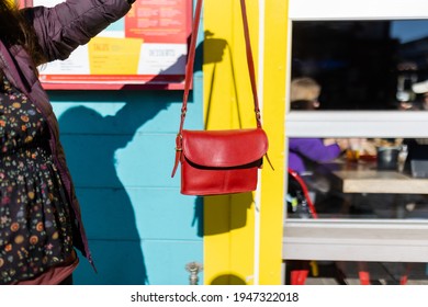 Woman Holding Cute Red Handbag Outside Yellow And Blue Building. Pretty And Small Purse With Long Strap Hanging Outside Restaurant Window On Sunny Day. Fashion And Accessories