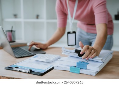 Woman holding document file stack of business document paper looking at laptop computer flicking through clients files on table, searching for and picking a file in home office.