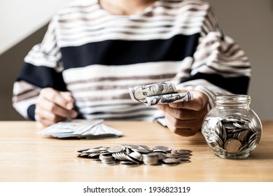 Woman Holding Dollar Bills, On The Table A Large Pile Of Coins And Banks, She Is Managing To Divide The Money To Save Money And Invest It To Make It Grow Even More. Saving Money And Investing Concept.