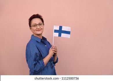 Woman Holding Finnish Flag. Nice Portrait Of Middle Aged Patriotic Lady 40 45 50 Years Old With A National Flag Of Finland Over Pink Wall Background. Learning Finnish. Travel To Finland Concept