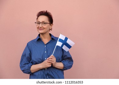 Woman Holding Finnish Flag. Portrait Of Middle Aged Woman 40 50 Years Old With A National Flag Of Finland Over Pastel Pink Wall Background.  Learning Finnish Language. Travel To Finland Concept.