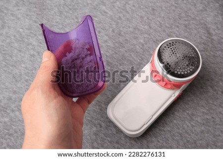 Woman holding fluff collector with lint of fabric shaver near light grey cloth, closeup