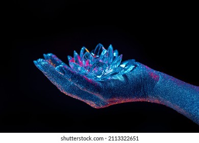 Woman holding fragile crystal lotus flower. Hand covered with shining glitter under neon colored light. Concept of religion, art, meditation, chakras, spiritual inner world