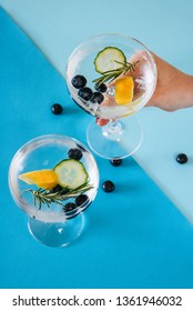 Woman Holding A Gin And Tonic Drink With Blueberries, Cucumber And Rosemary On A Blue Geometric, Colorful Background.