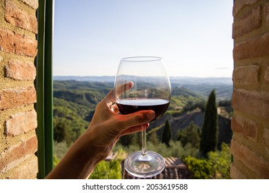 Woman holding a glass of red wine with beautiful landscape of Italy in a background on a sunny day. View frm the window.