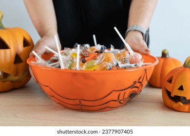 Woman holding a Halloween candy bowl filled with sweets for trick-or-treating. Female hands and Halloween-themed decorative treat bowl container. Jack-o'-lantern pumpkins placed around the table. - Powered by Shutterstock