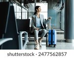 Woman is holding handle of blue suitcase and looking around airport terminal. Business woman sitting in airport and waiting for starting her travel voyage