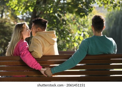 Woman holding hands with another man behind her boyfriend's back on bench in park. Love triangle