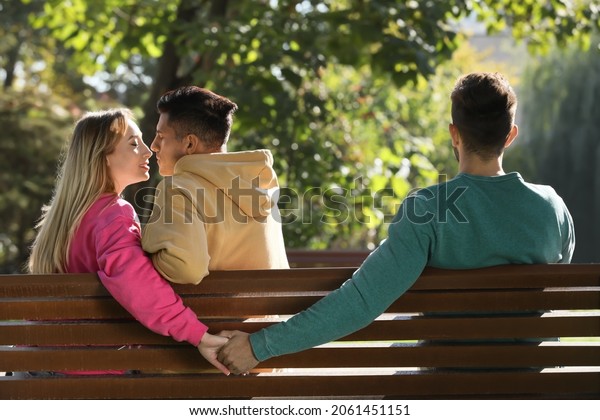 Woman holding hands with another
man behind her boyfriend's back on bench in park. Love
triangle