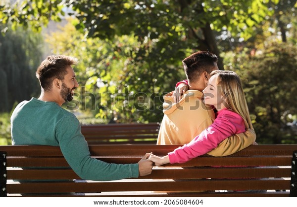 Woman holding hands with another\
man behind her boyfriend\'s back on bench in park. Love\
triangle