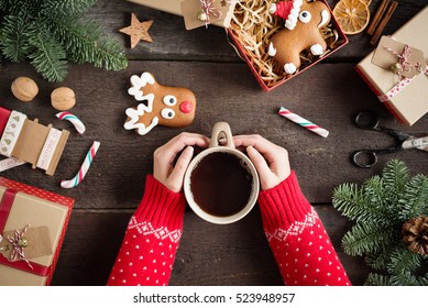 Woman holding in hands hot christmas tea with candy cane against decorations, gift boxes, ribbon and ginger bread on wooden board. Xmas concept. Eye bird view. 