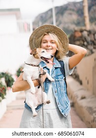 Woman holding hat and carrying small dog looking at camera in the street.