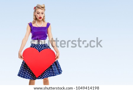 Woman holding heart symbol, dressed in pin-up style dress with polka dot, with copyspace area for slogan or advertising text message, on blue background. Blond model in retro fashion concept.