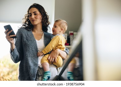 Woman Holding Her Baby And Texting On Her Phone. Female With Her New Born Baby Using Mobile Phone At Home.