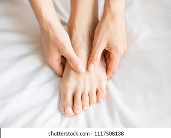 Woman holding her foot on bed with white sheets. - Shutterstock ID 1117508138