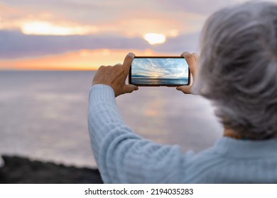 Woman Holding Her Mobile Phone Taking A Picture Of The Sunset Over The Sea, Dramatic Sky And Sunlight