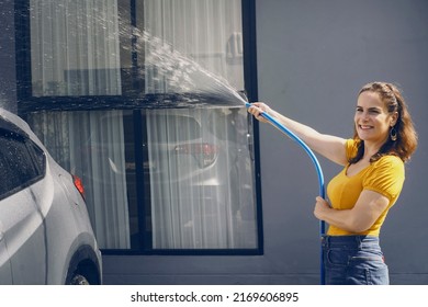 Woman Holding A Hose Using Water To Wash A Dirty, Dusty Car In Front Of The Garage At Home