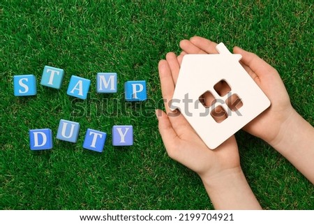 Woman holding house figure and blue wooden cubes with text Stamp Duty on green grass, top view