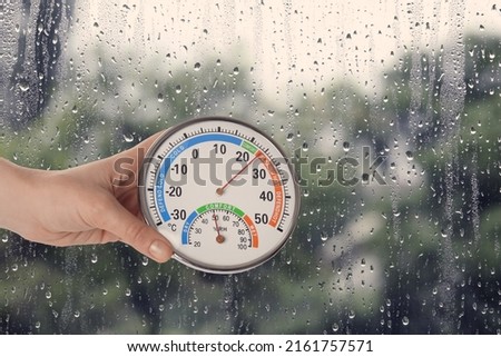Woman holding hygrometer with thermometer near window on rainy day, closeup