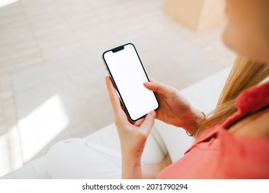 Woman holding iPhone 13 pro with a white screen mock up, resting on a sofa in living room at home. Rostov-on-Don, Russia - November 1 2021