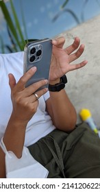 Woman Holding An Iphone Cell Phone In Gray Color, Sitting, Relaxing On A Beach. Iphone With A Transparent Protective Cover. Malang, Indonesia - 02.11.2022