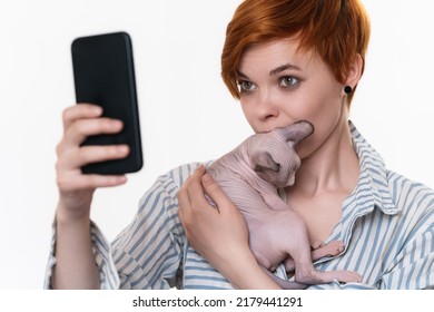 Woman Holding And Kissing Kitten, Taking Selfie Self Portrait Photos On Smartphone. Young Adult Redhead Hipster Dressed Striped White-blue Shirt. Part Of Series. Studio Shot On White Background.