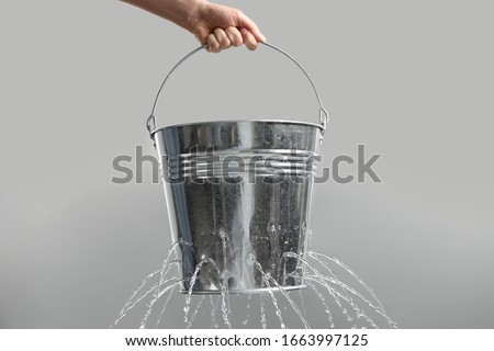 Woman holding leaky bucket with water on light grey background, closeup