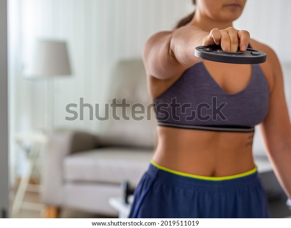 Woman holding a\
lightweight disk for working out arms and triceps at home.\
Exercising in living room for weight loss and losing body fat.\
Training at home online\
class.