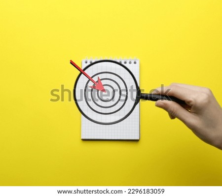 Woman holding magnifying glass over notebook on yellow background, top view. Illustration of target and arrow