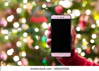 Woman Holding Mobile Phone And Showing Screen With Christmas Tree Lights On The Background. Mockup For Greeting. Concept.  